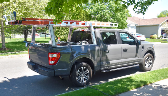 Gray pickup truck with ladders on a ladder rack and equipment in the bed parked by a road curb in a neighborhood